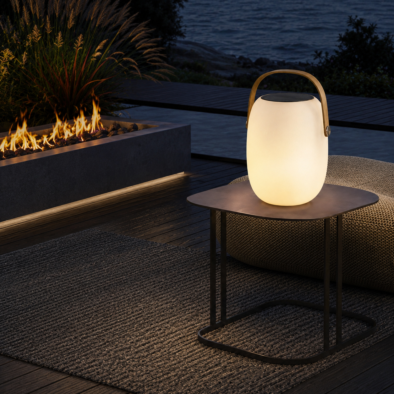 Aire portable outdoor lantern on metal side table beside fire pit and woven rug overlooking water at night