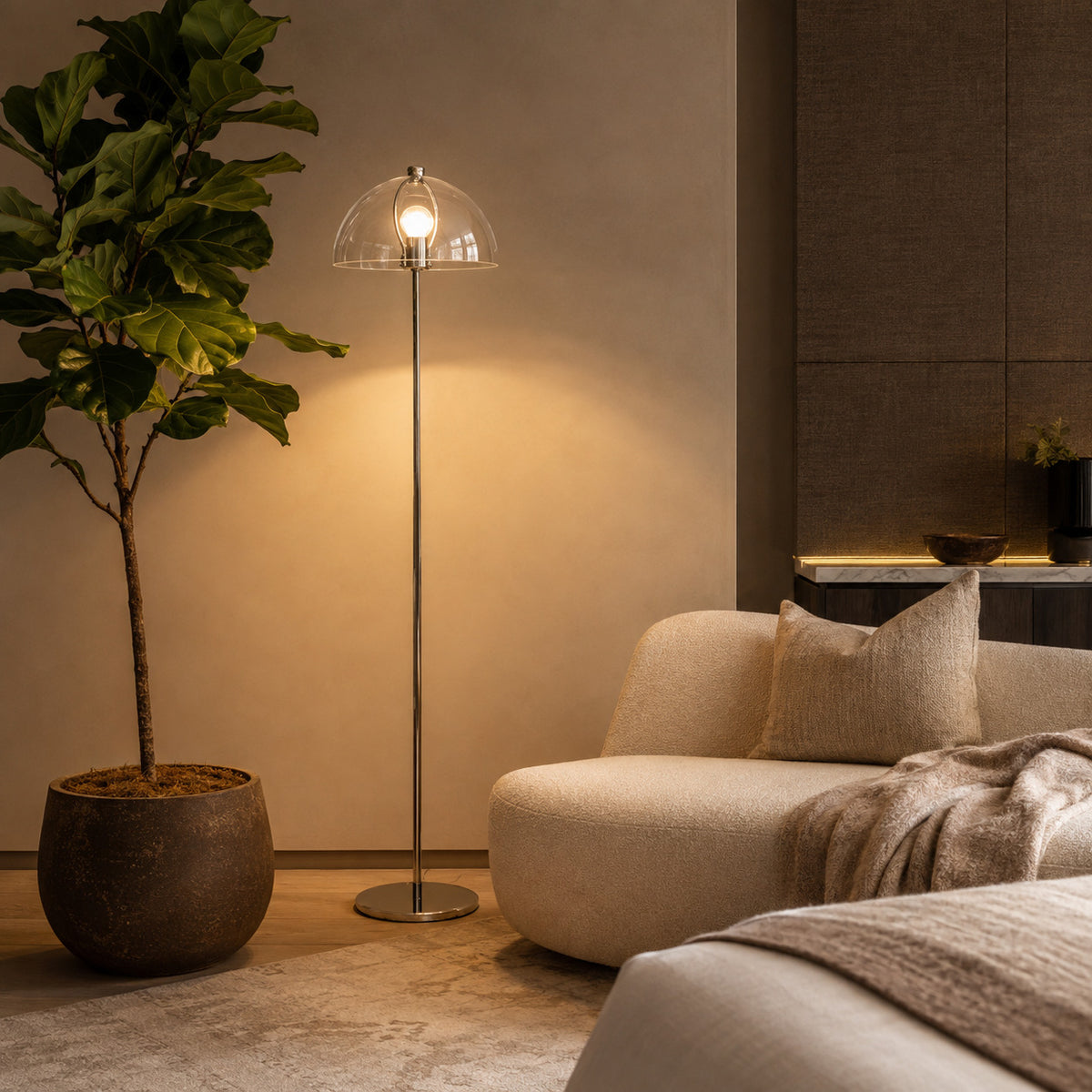 Benavente floor lamp in clear acrylic, lit beside a potted fig and a pale sofa