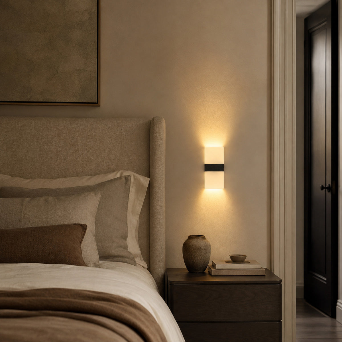 Anços sconce in black with warm white light beside a bed, washing light up and down the wall