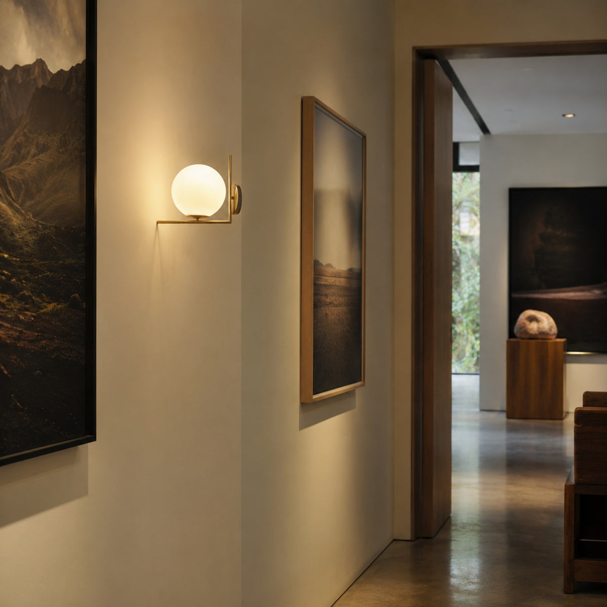 Ceira globe wall sconce in brass, lit on a hallway wall beside framed art