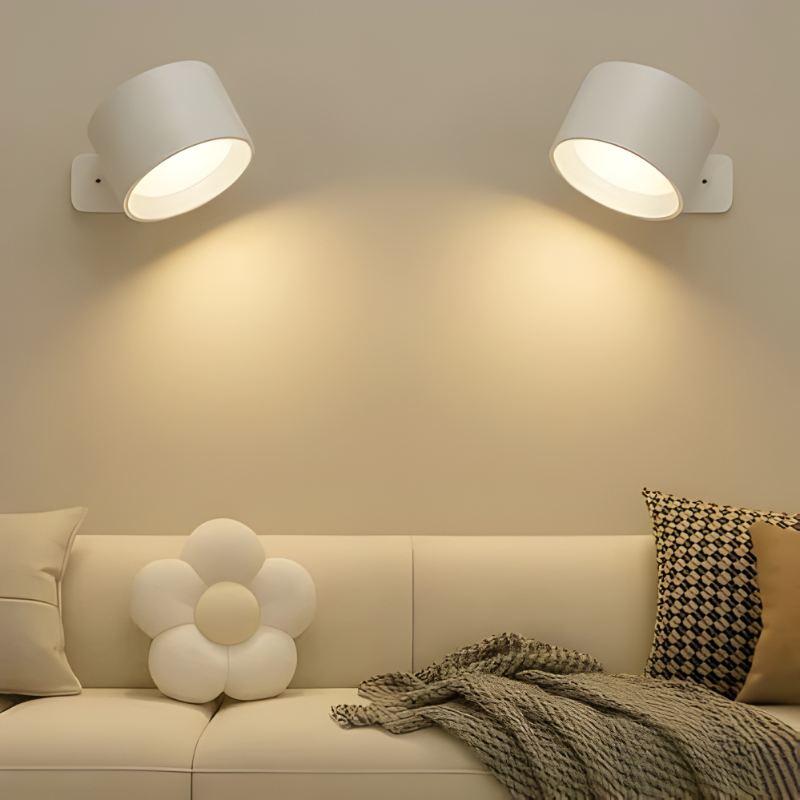 Two Nisa wall sconces, white, lighting a living room wall above a sofa with a flower cushion.